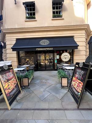 From sidewalk facing venue. Path ahead to entrance at back. A-boards on left and right. Planters behind boards. Outdoor seating on either sides.