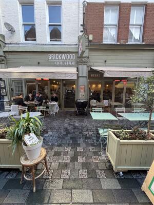 From sidewalk facing venue. Opening ahead between planters. Path behind between outdoors seating to entrance on back.