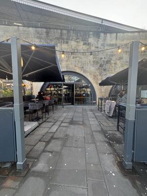 From sidewalk facing venue. Path ahead to entrance at back. Outdoor seating on both sides of path.