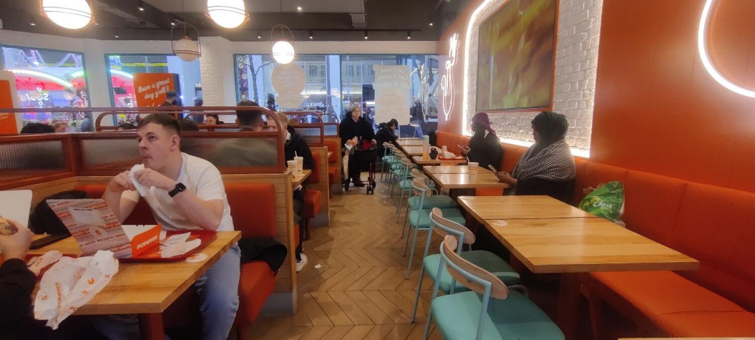 From drink dispenser facing left. Booth on left. Path to more seating in center. Table, chairs, half booth along right.