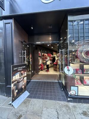 From sidewalk facing venue. Archway ahead. Signboard on left. Glass doors with glass frames at back. Pull from center to open.