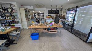 From entrance facing straight. Product shelves with along left. Table with bread and snacks in centre. Counter behind. Fridges along right.