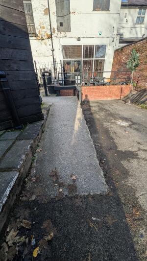 From back of venue facing venue. Concrete ramp leading up to entrance doors ahead. Brick wall with railing on right of ramp.