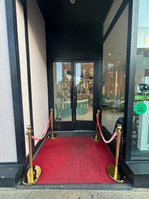 From sidewalk facing venue. Black framed archway ahead. Red queue cords after on both sides.