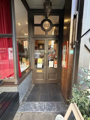 From sidewalk facing venue archway ahead. Bench on left. Signboard on right. Glass door with brown frames at back. Pull from center to open.