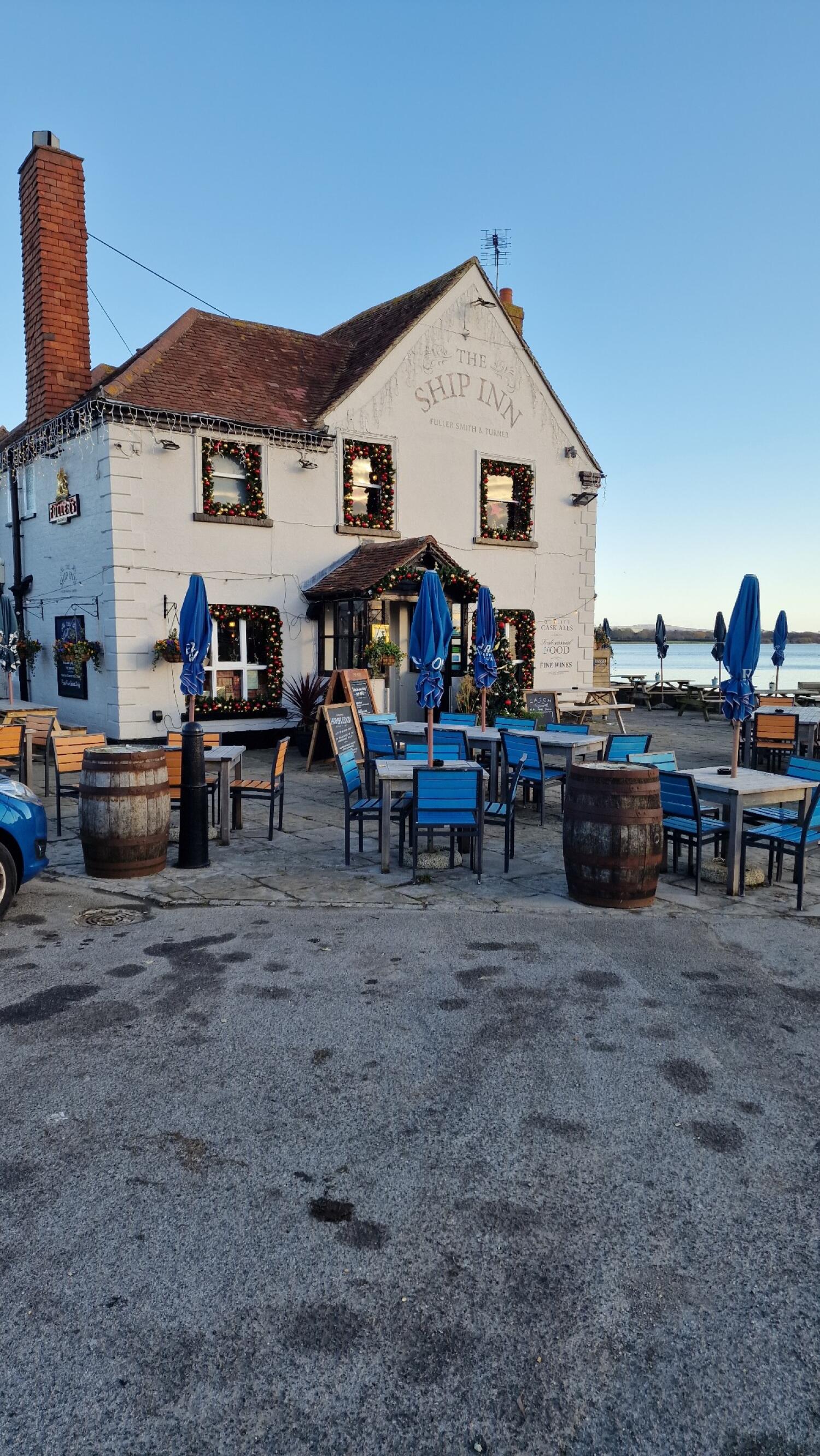 Tables and chairs with blue parasols in front. Entrance door at centre of building. Additional seating and barrels to right near waterfront.