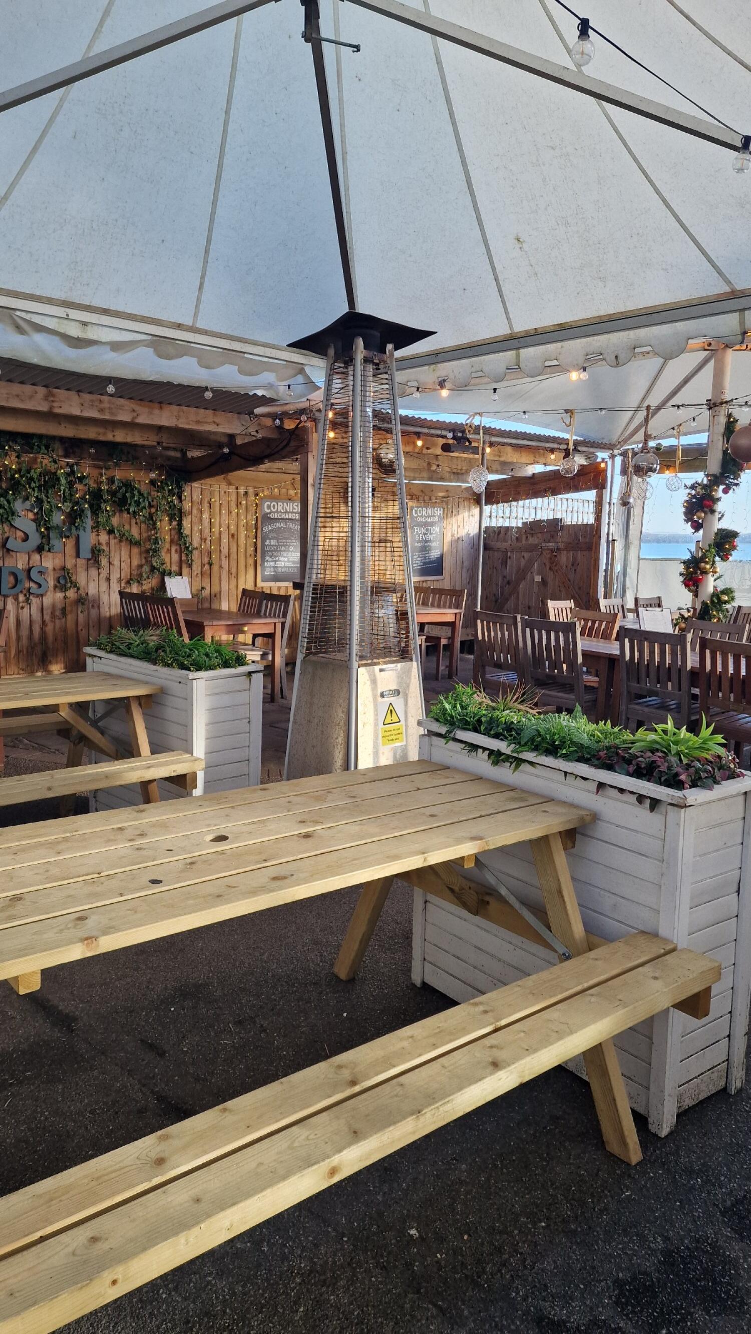 Wooden picnic benches in front. Planters with greenery on each side. Wooden tables and chairs behind, under large white tent.