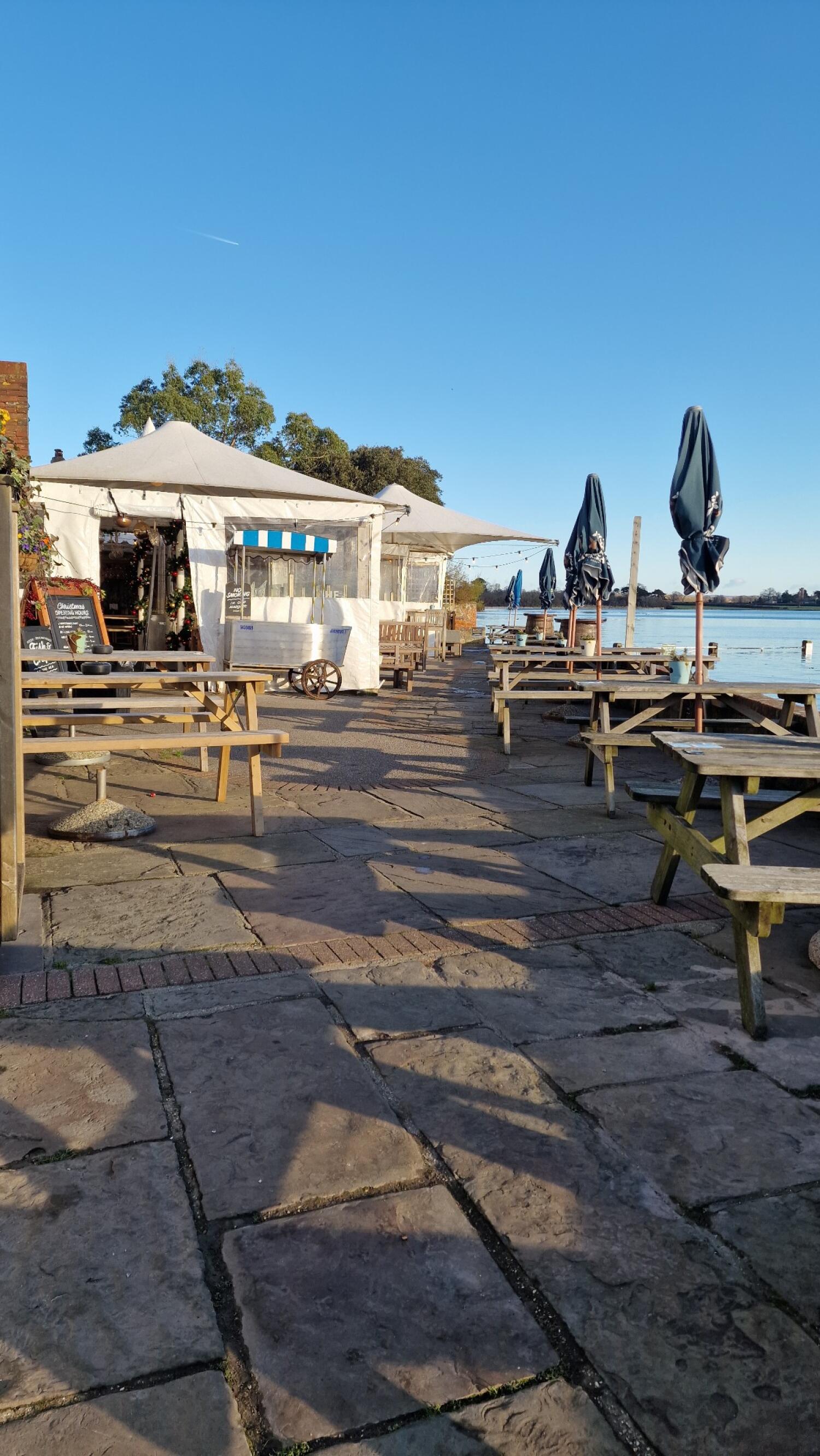 Picnic benches along left and right. Path leads to tent ahead. Closed parasols beside benches on right by water.