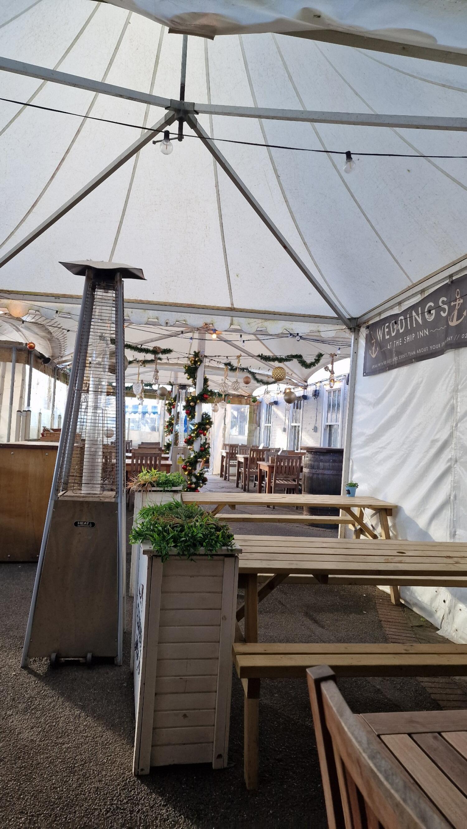 Wooden tables and chairs along left and right. Planter with greenery in centre. Tent covering entire space.
