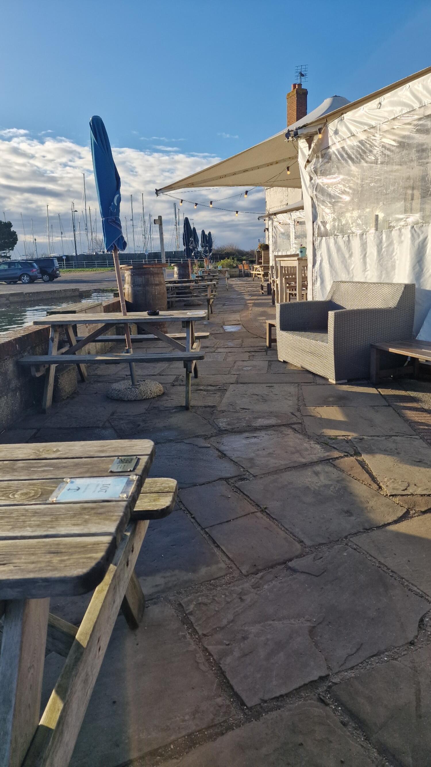 Picnic tables with closed blue parasols along left. Wicker armchair and wooden benches along right. Pathway continues ahead beside waterfront.
