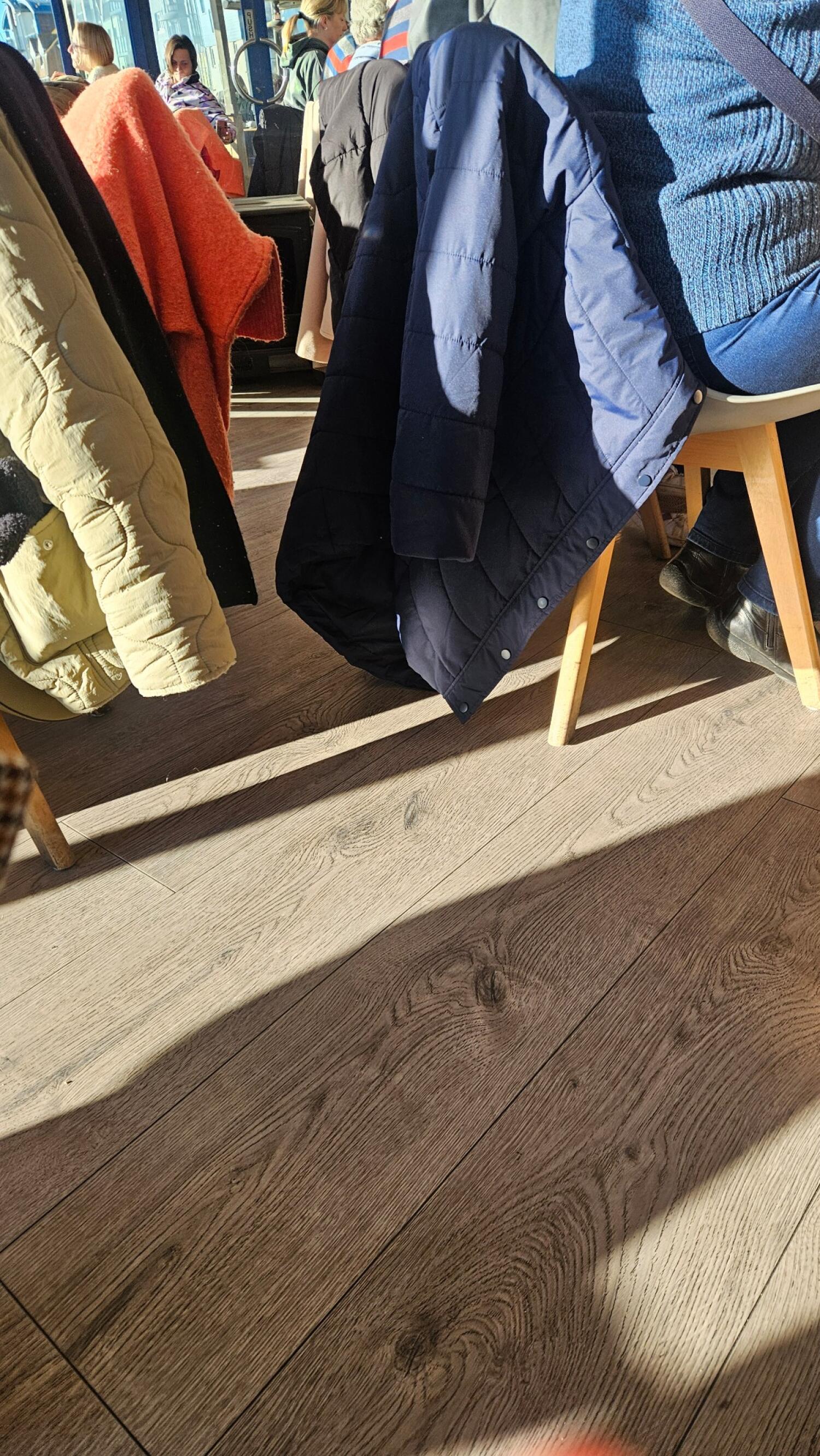 Tables and chairs closely arranged. Coats draped over chair backs. Bright sunlight streaming across space.