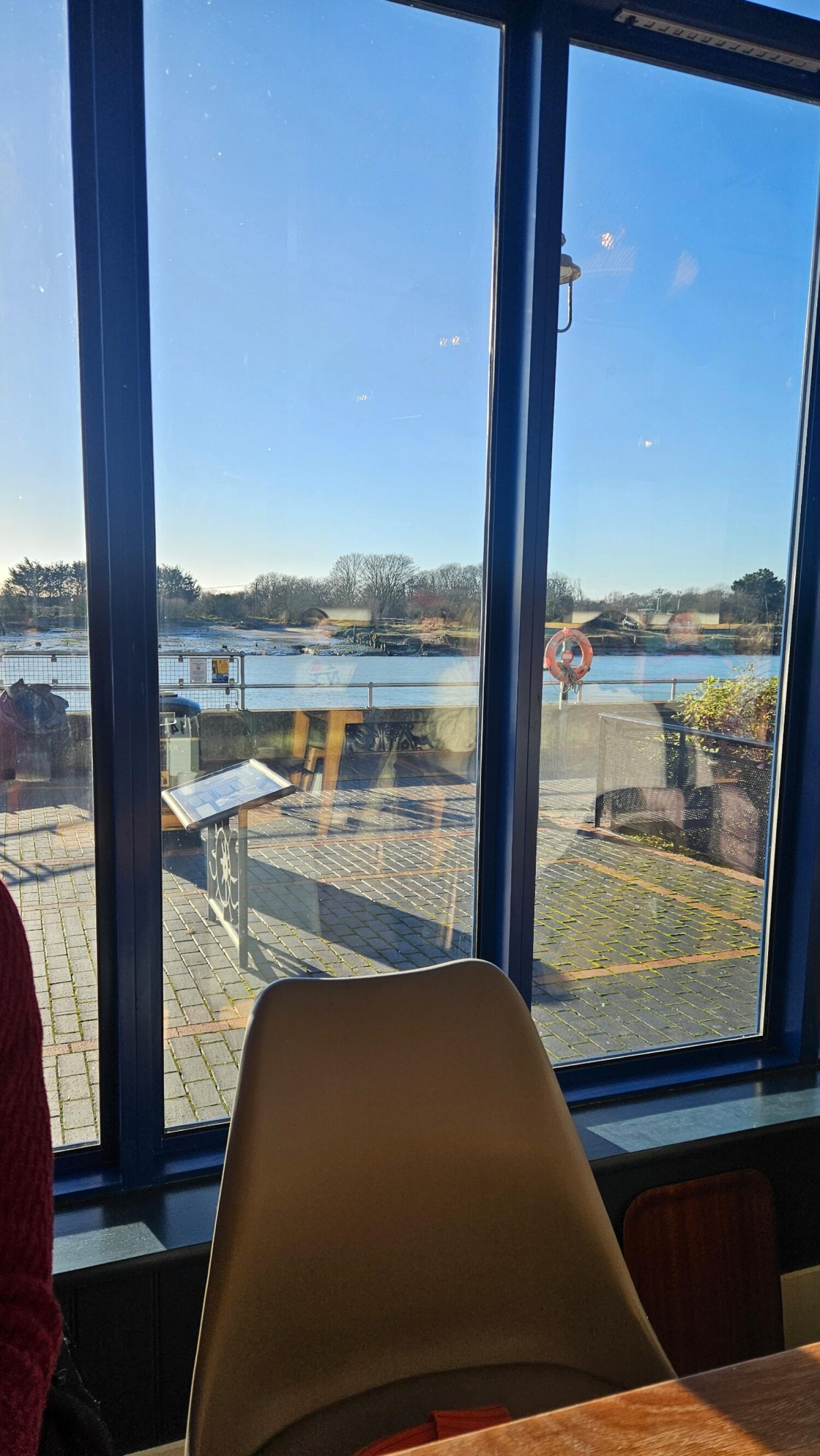 Chair in foreground facing large blue-framed windows. Outdoor paved area beyond windows. River and trees visible in background.
