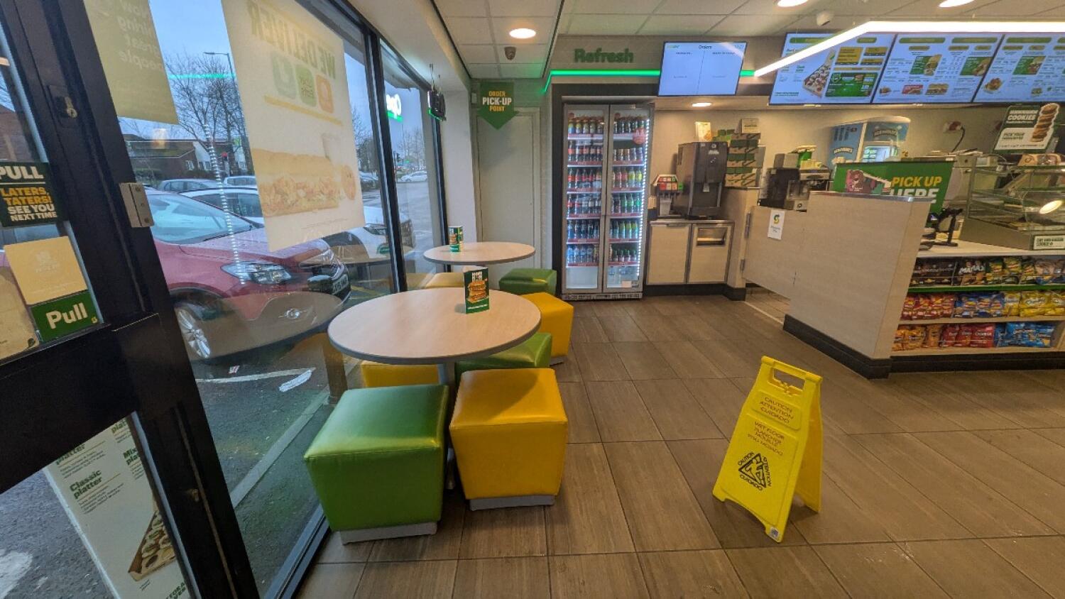 From entrance facing left. Tables with cube stools along left wall. Fridge and drinks station at back. Counter with crisps on right.