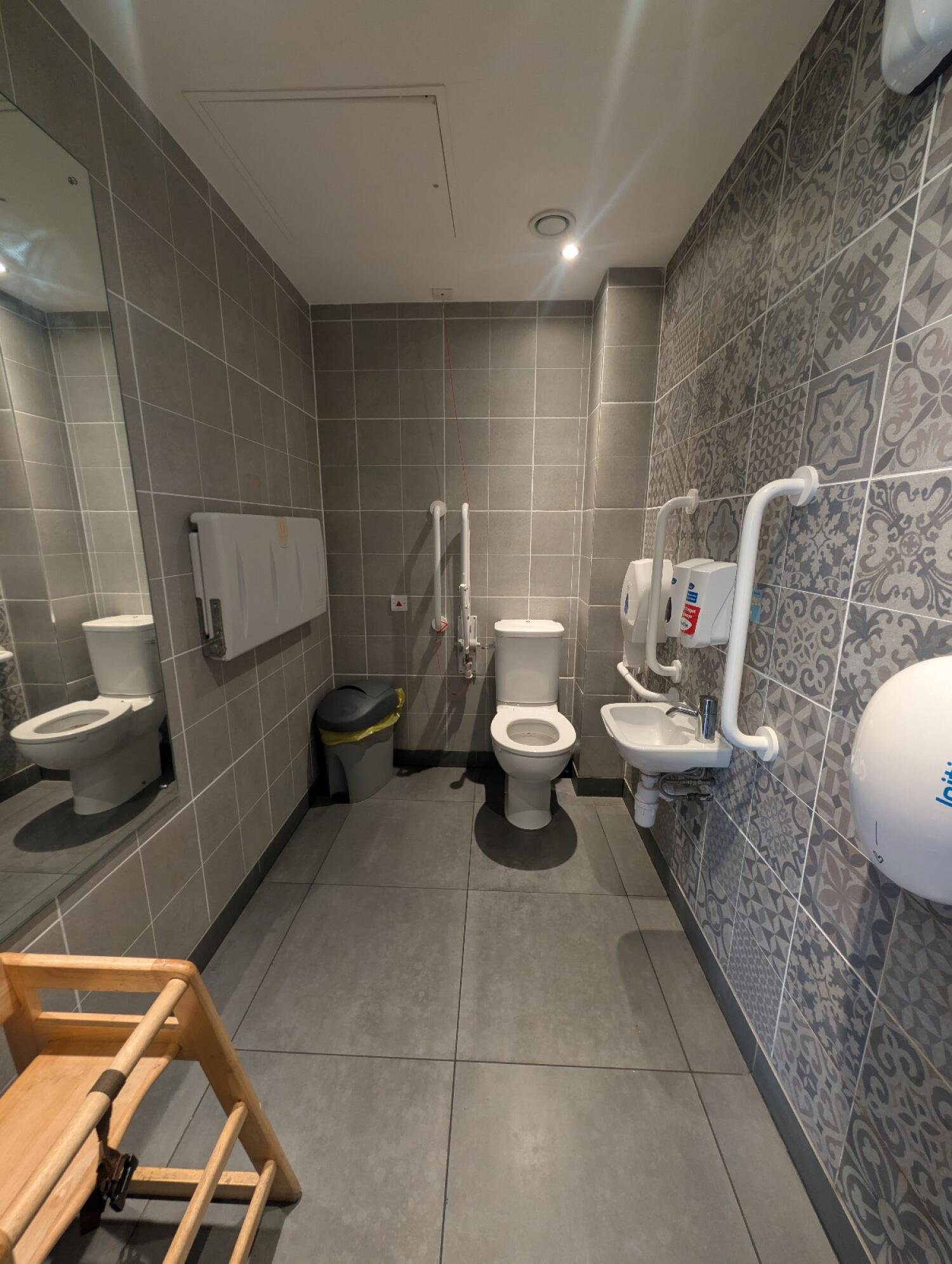 From bathroom door facing straight. Sink on right wall. Toilet at back right. Pull cord to it's left. Baby change table on left wall.