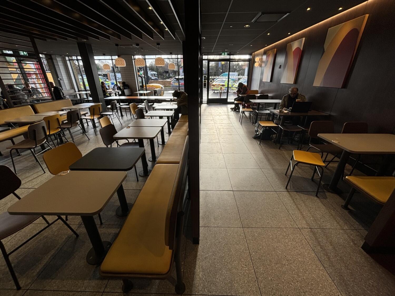 From end of kiosks facing left. Divider ahead. Half booths and chair along left. Tables and chairs along right wall.