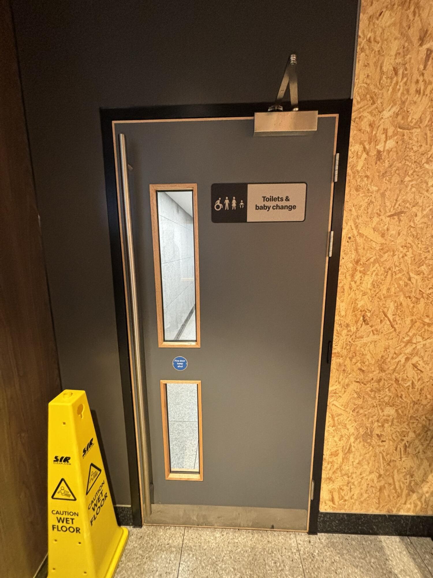 From end of side space seating facing left. Grey preceeding toilet door ahead. Pull handle on left to open.