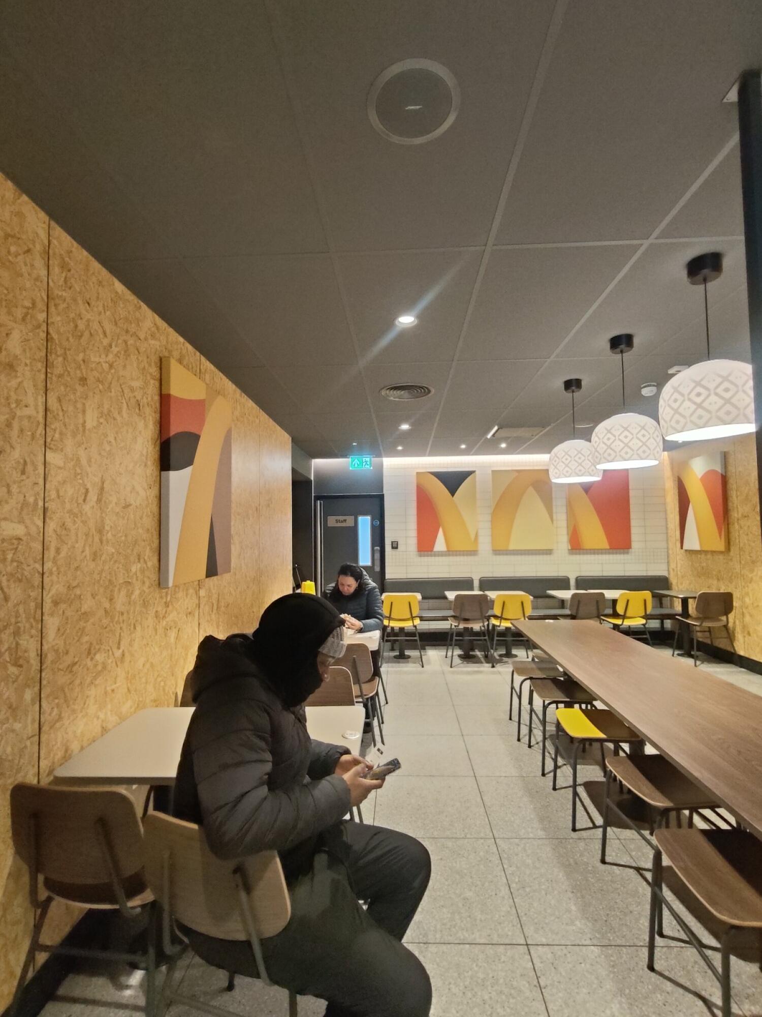 From left end of partition facing straight. Tables and chairs along left wall. Table and stools in centre. Half booths at back.