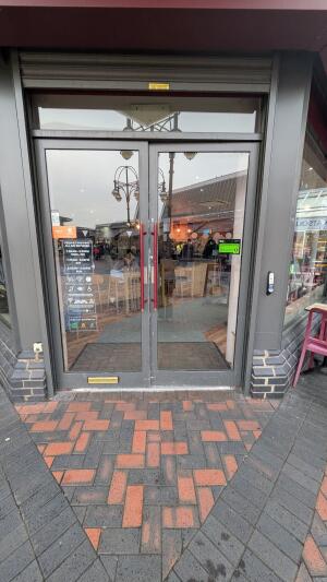 Grey metal-framed glass doors. Push from red handles at centre to open. Doorbell for assistance on right of door.