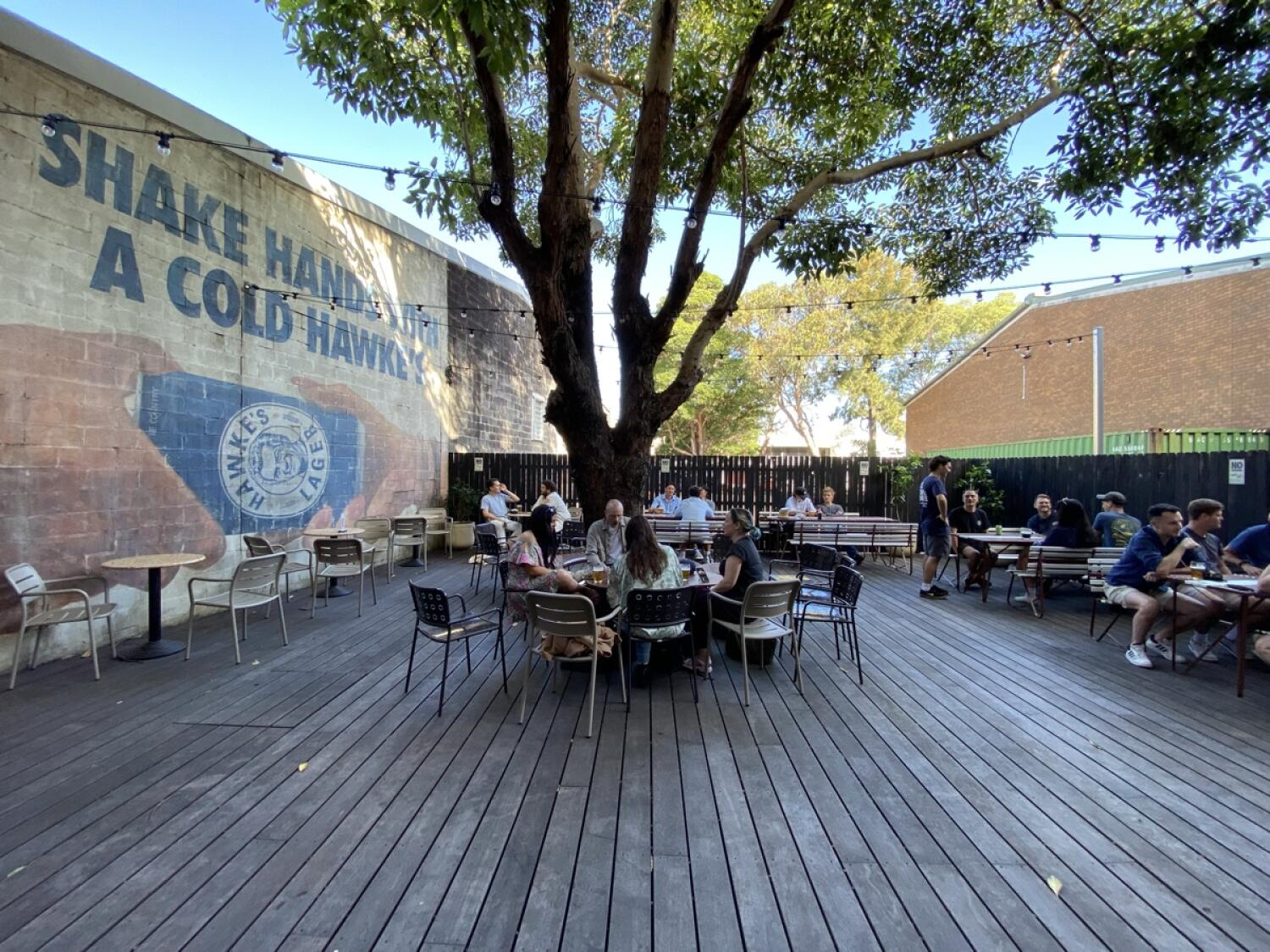From door facing straight. Tables and chairs ahead. Large tree in centre with seating around. Long tables with benches along back of tree.