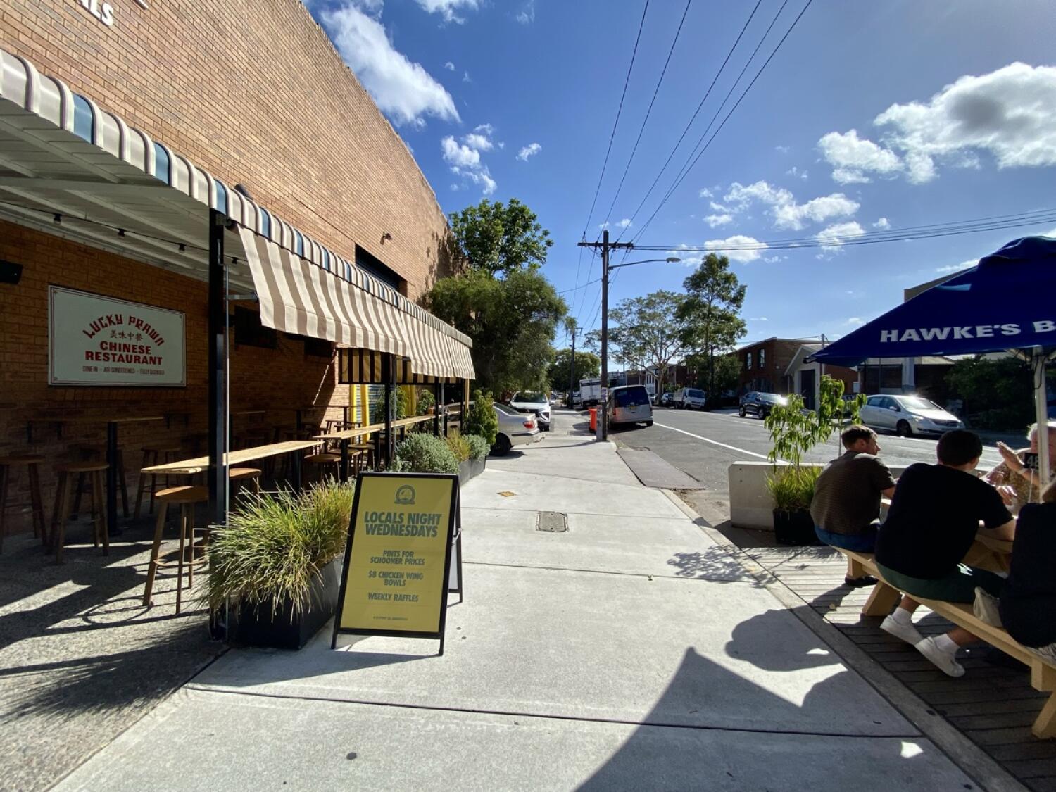 From entrance facing right. Path ahead. A-frame sign on front left. Wooden picnic tables with parasols along right.