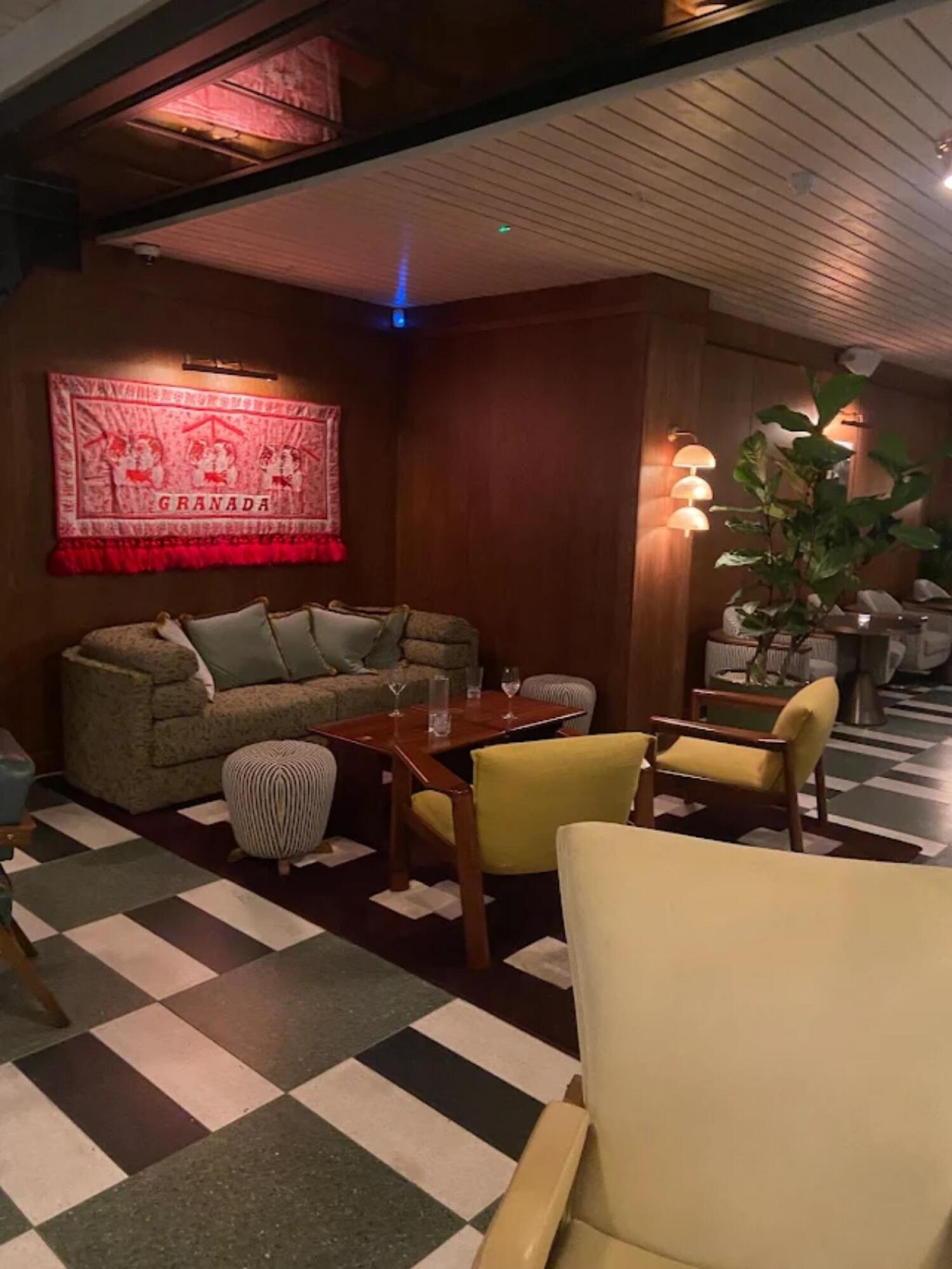 Sofa along left wall with two footstools in front. Wooden table with two yellow chairs at centre. Plant and additional seating along right wall.