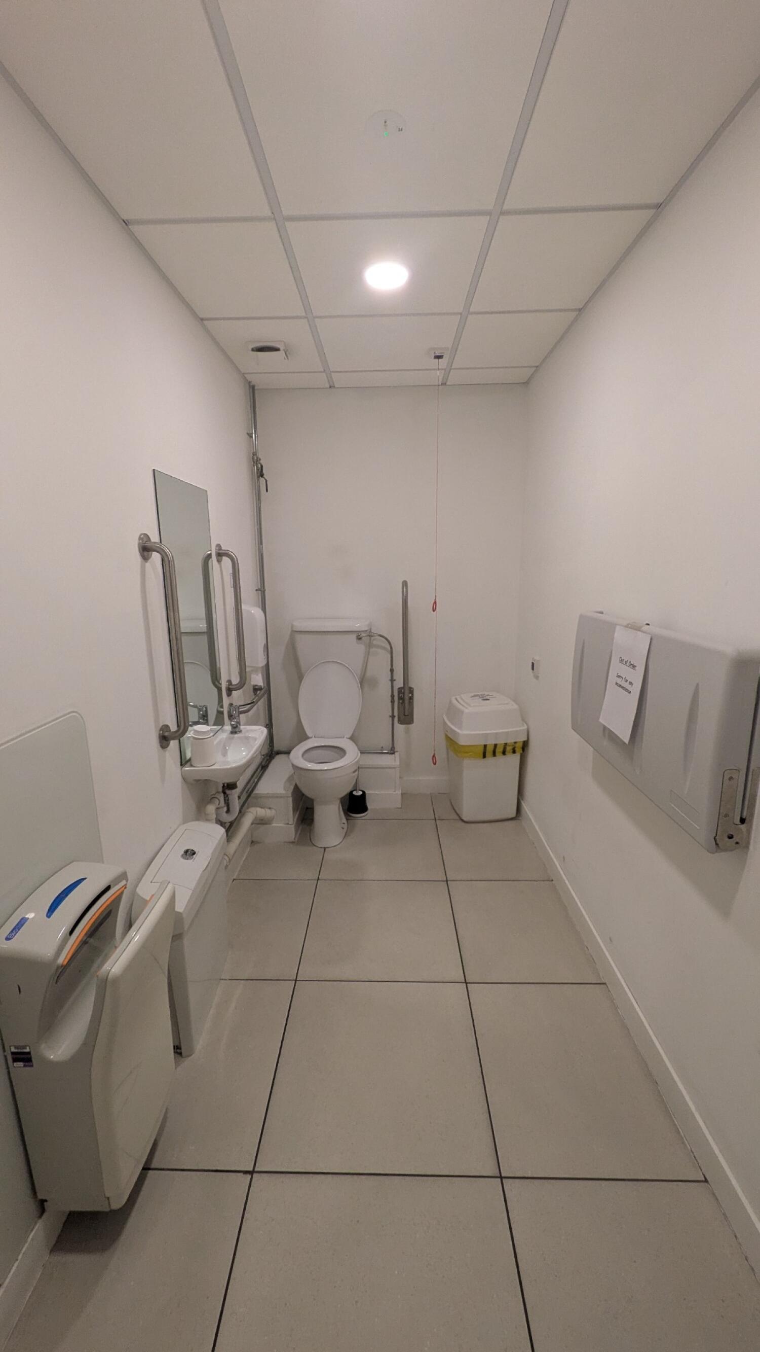 From door facing straight. Sink on left wall. Toilet at back wall. Emergency pull cord to its right. Baby changing station on right wall.
