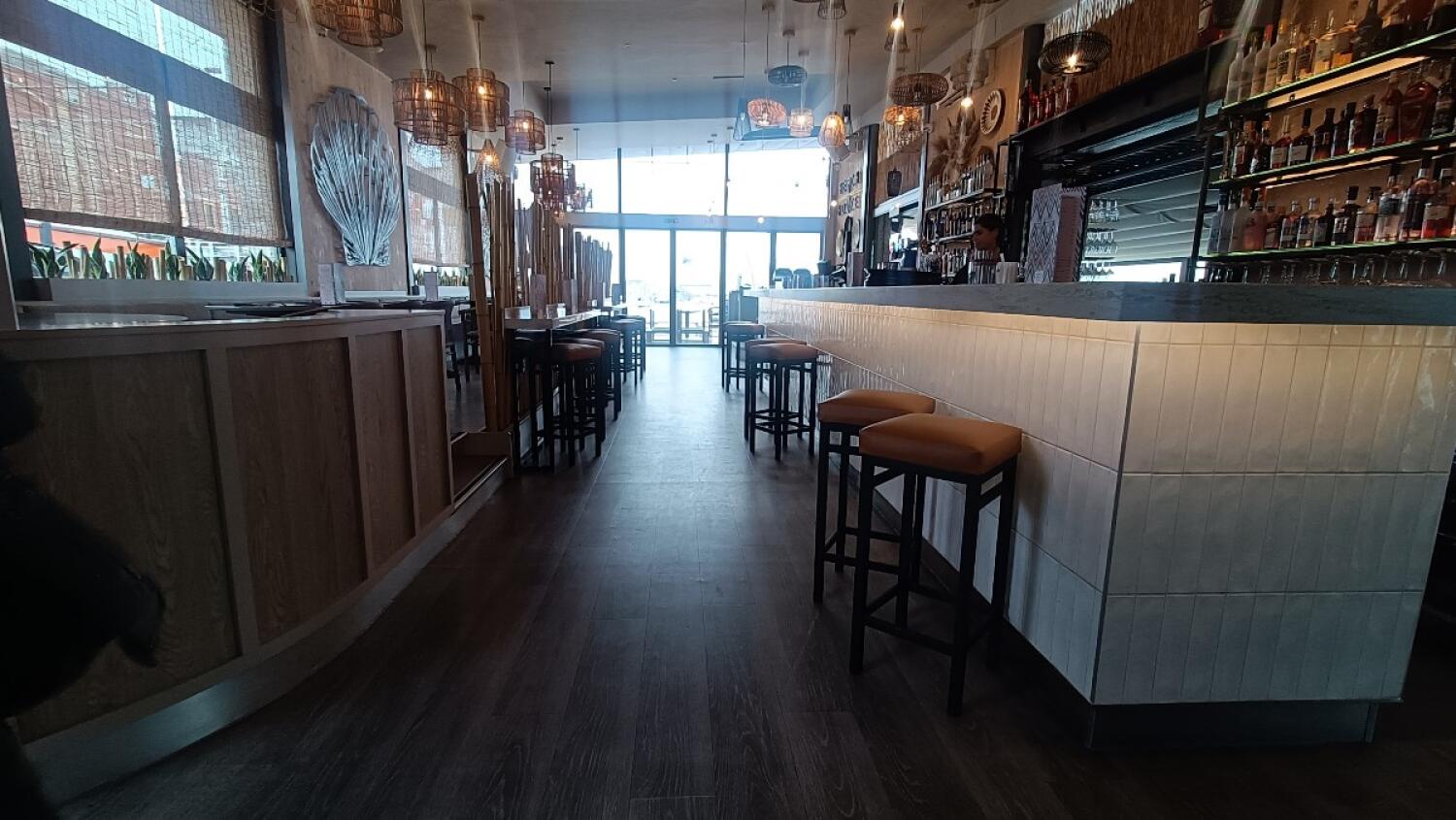 From welcome desk facing left. Partition along left. High table and stools along it at back left. Bar counter with stools along right.