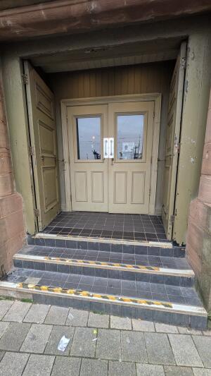Glass doors with green frame. Push in center to open. Three steps up before door.