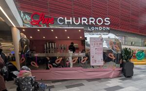 From mall facing venue. Pink and silver churros kiosk with serving counter at centre. Menu board displayed on right side.