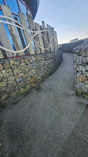 Path curves right between stone-filled wire fences. Building wall along left side of path.