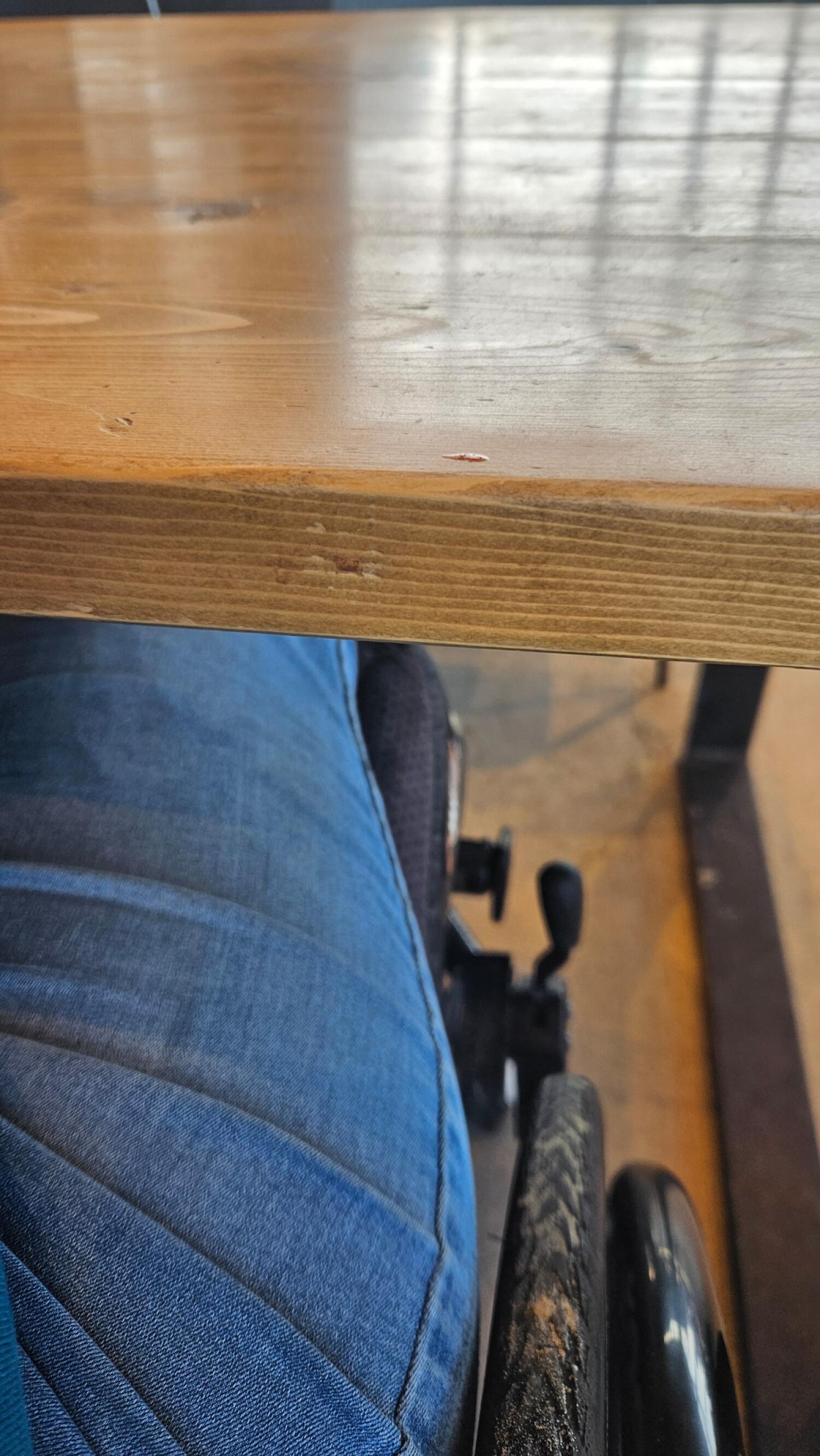 Close view of wooden table edge. Person in wheelchair seated underneath.