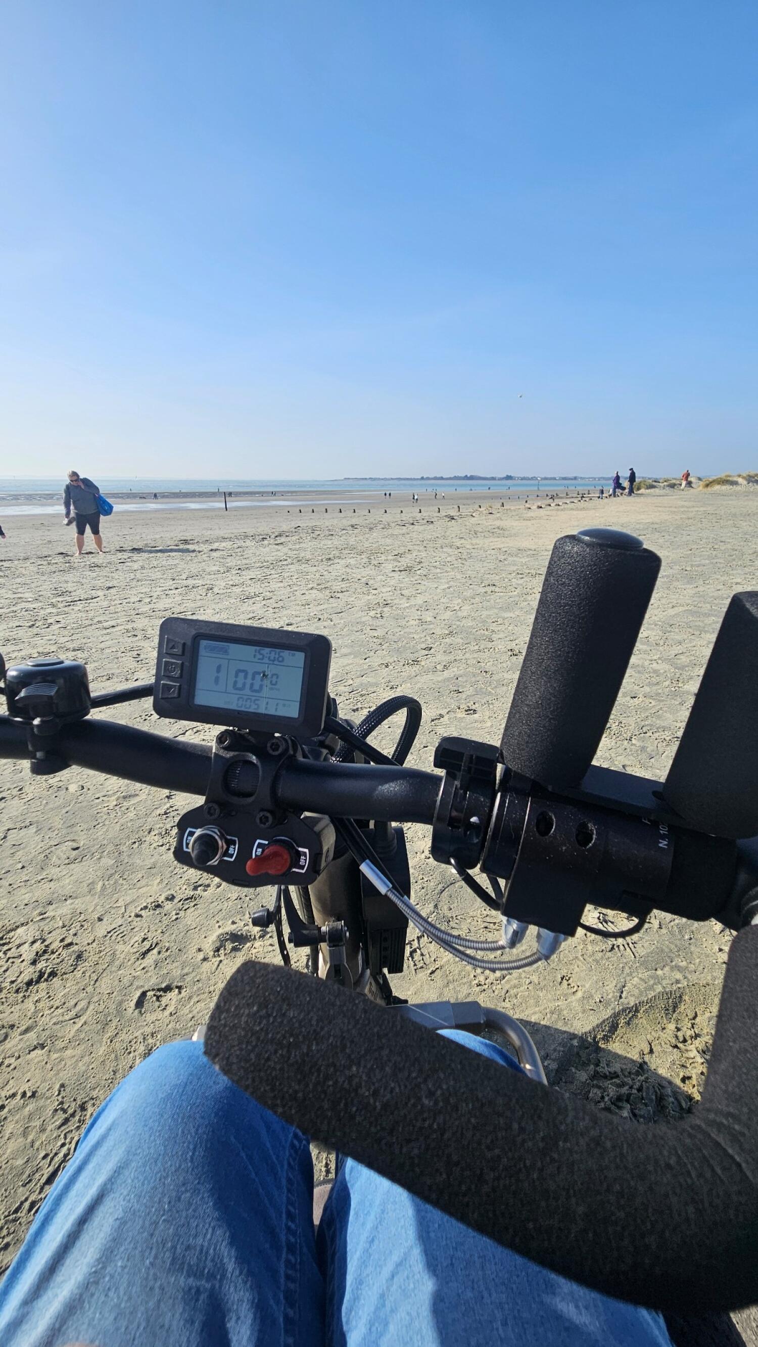 View from seated position on mobility scooter facing sandy beach. Scooter handlebars and digital display visible in foreground. People walking and sitting near shoreline in background.