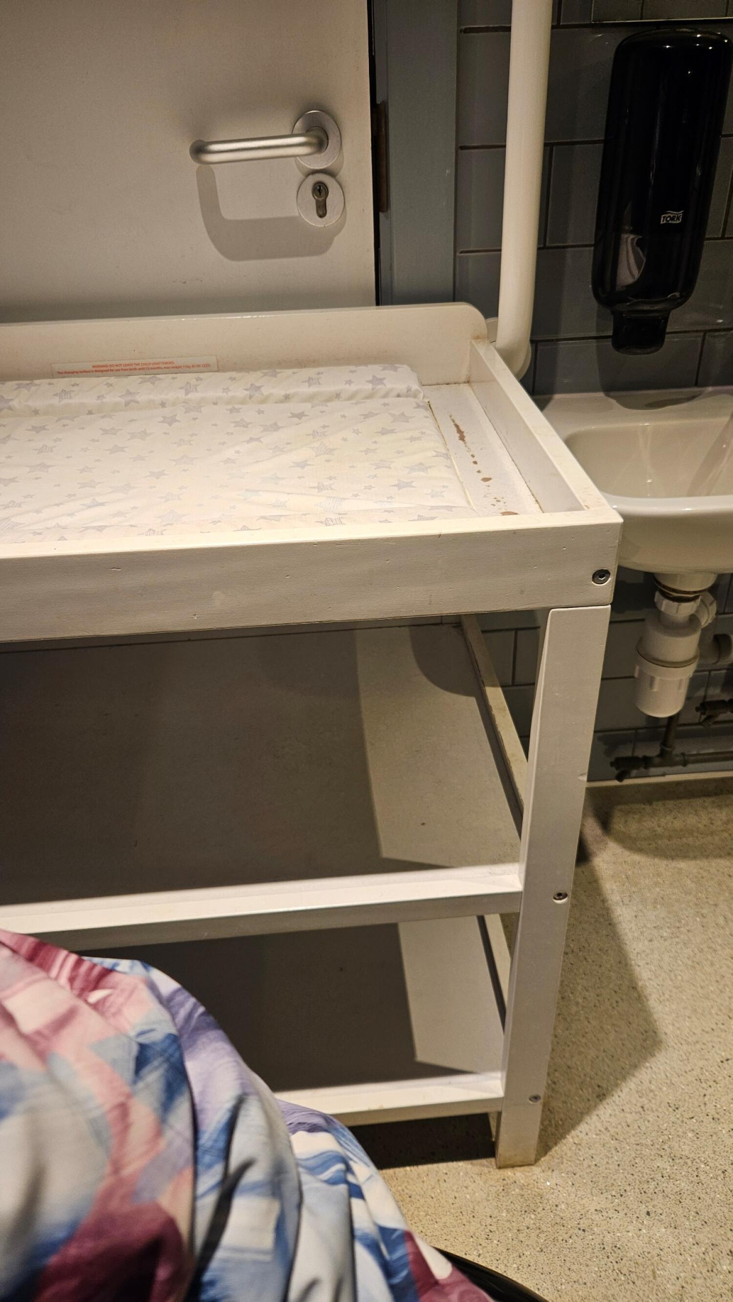 Baby changing station in centre. Sink on right wall. Door behind with handle on left.