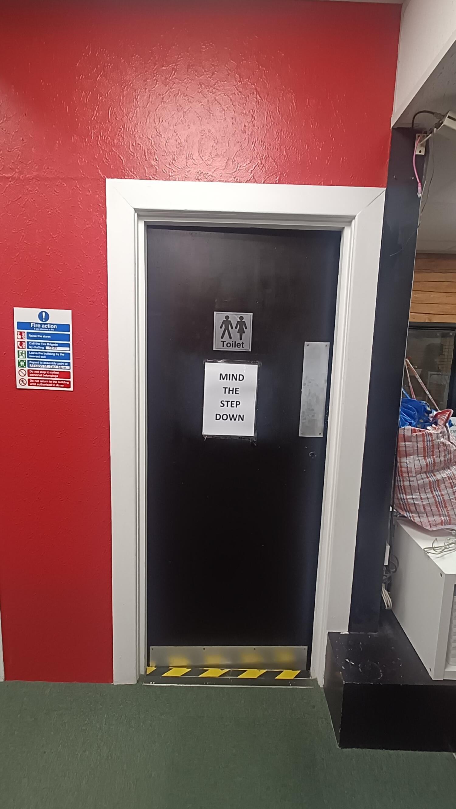 From open space facing left. Black common toilet door with white frame ahead. Push metal strip on right to open.