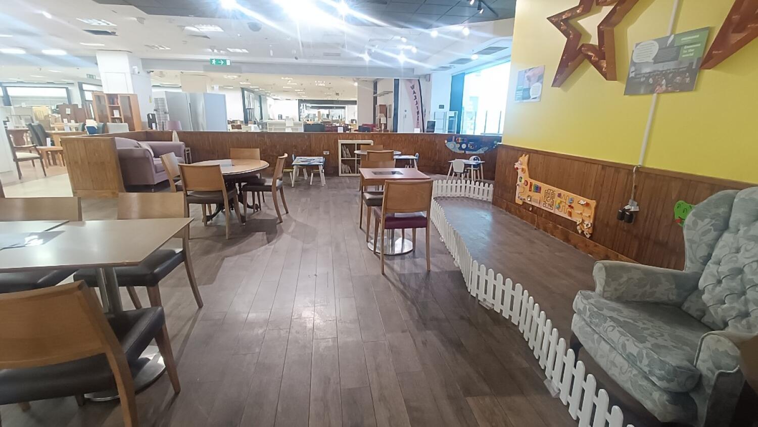 From second partition facing straight. Tables and chairs along left and centre. Couch and table at back left. Small fenced play area on right.