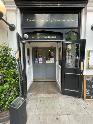 Wooden double doors with glass panels, handle in centre. Pull to open. Display board at right, plants at left of entrance.