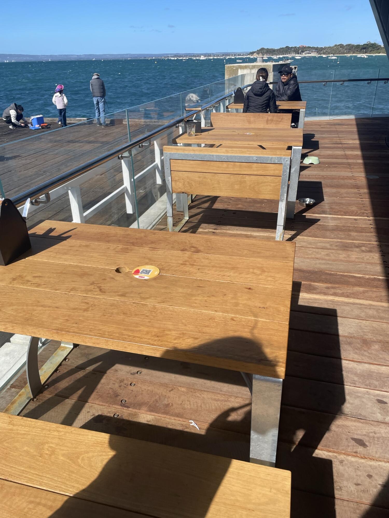 Wooden tables with benches along right side of deck. Glass railing with handrail along left. Sea and pier visible beyond railing.