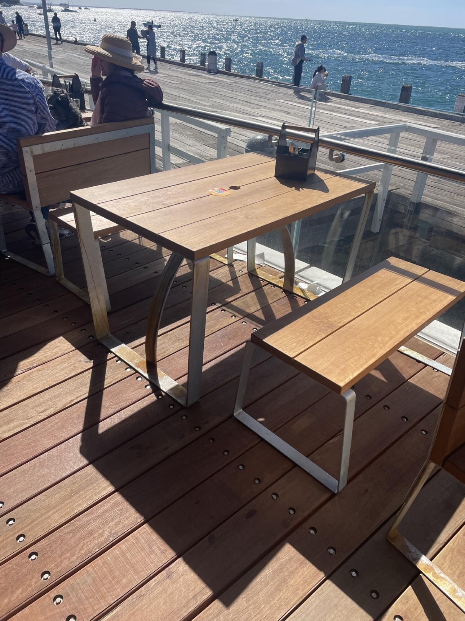 Bench seating along left. Wooden table with bench in centre. Ocean view beyond railing on right.