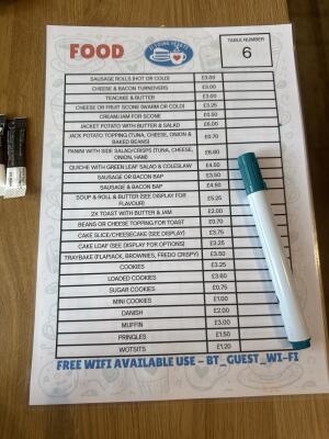 The image shows a printed food menu on a wooden table with a whiteboard marker placed on it. The menu is titled "FOOD" in red text and features the "2 Young Hearts" logo at the top, along with a table number "6" box. It lists various items such as sausage rolls, cheese & bacon turnovers, jacket pota
