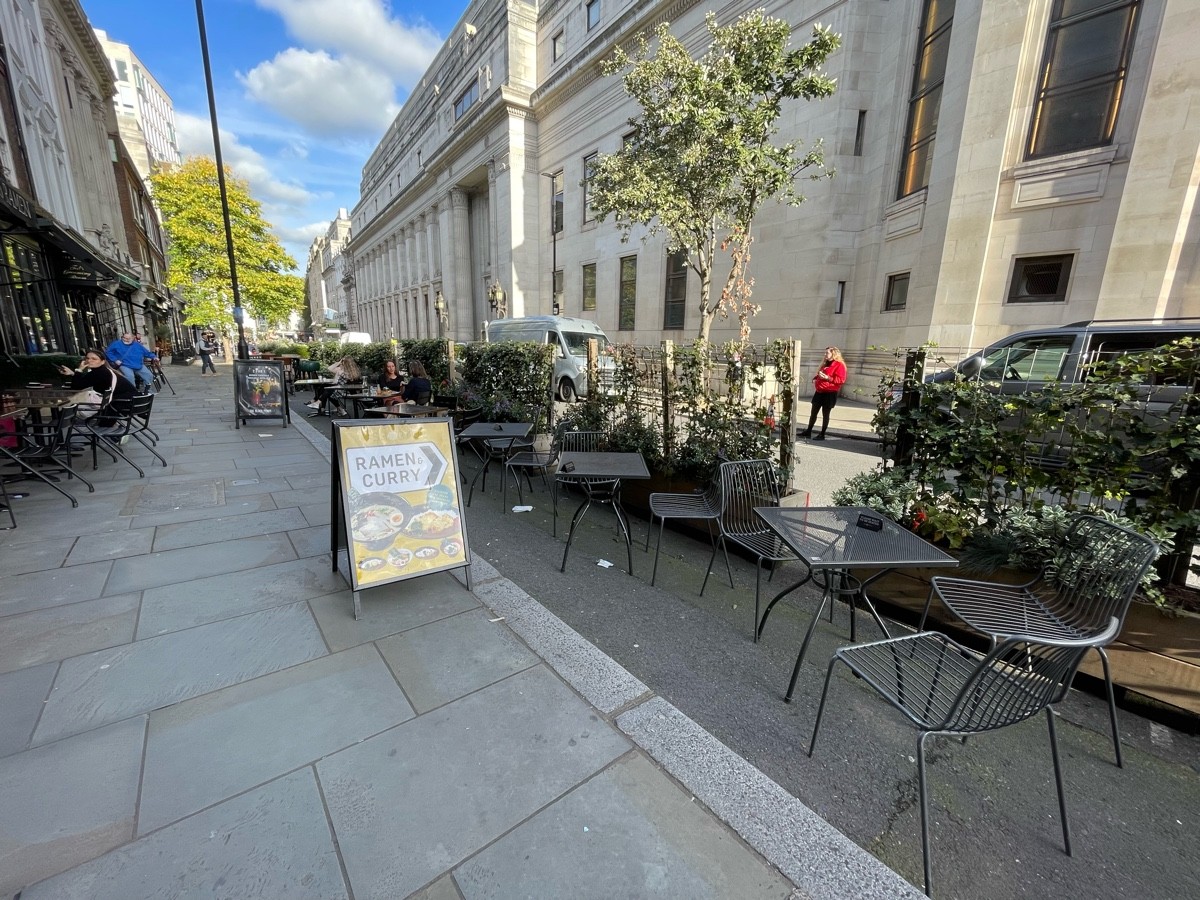 From outside entrance facing right. Pavement ahead, going straight. One step down from pavement to road. Tables along right. Planters along far right.