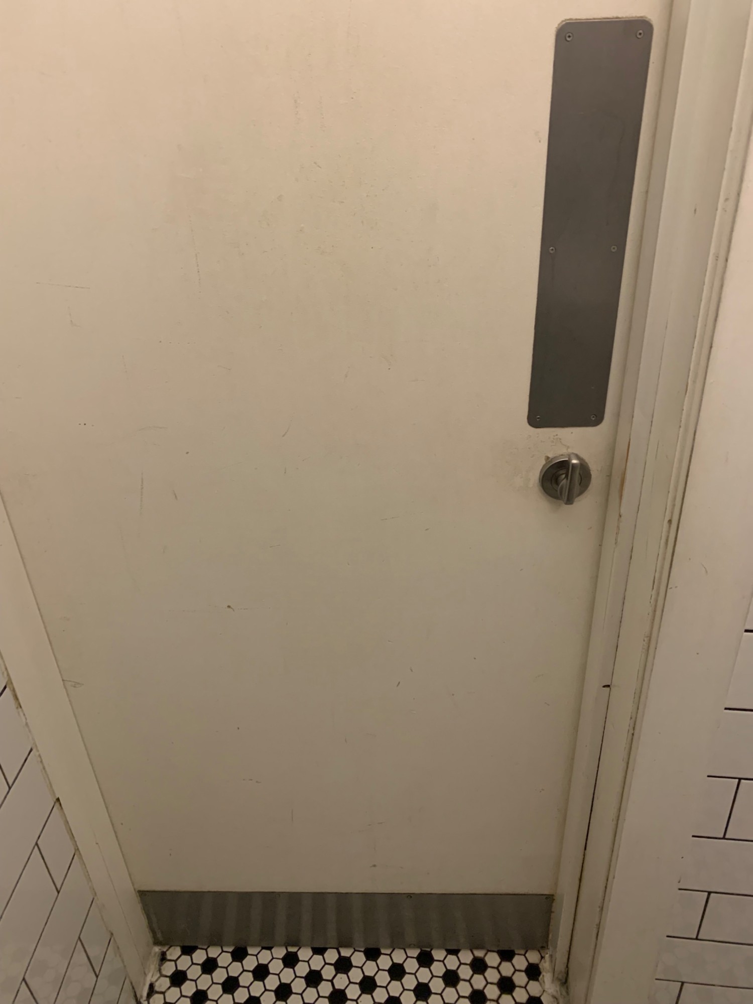 From sink, looking back. Back of toilet door ahead. White door with silver metal panel on right of door. Push to open.