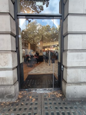Facing single glass door. Push silver vertical handle on right to open. Doorbell for assistance on right of door.