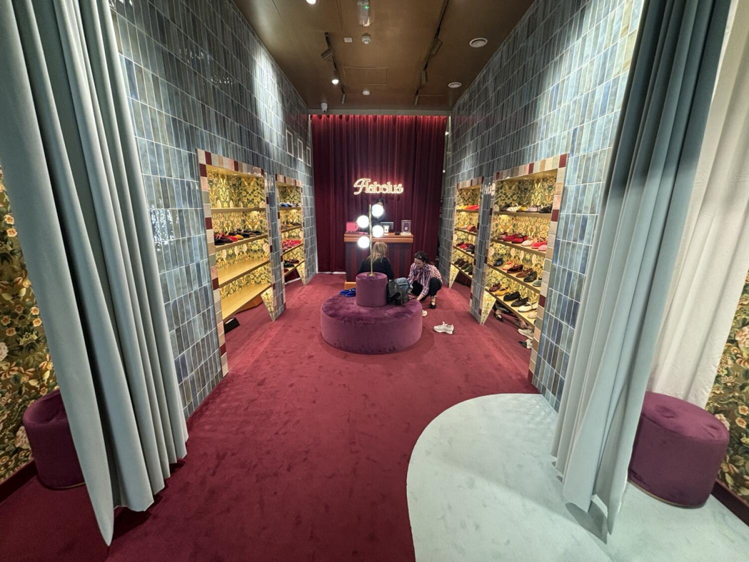 From opening facing straight. Fitting rooms on both sides. Product shelves at back left and right walls. Circular ottoman in centre. Counter behind.