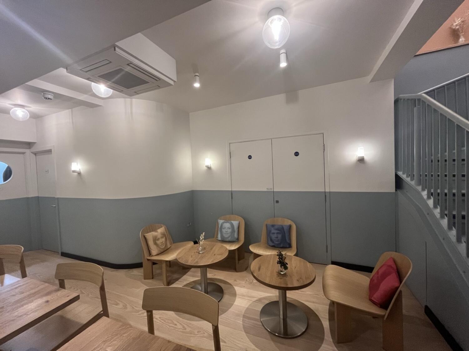From start of half booth facing right. Tables and chairs along left. Round tables and chairs in centre ahead. Staircase on right.