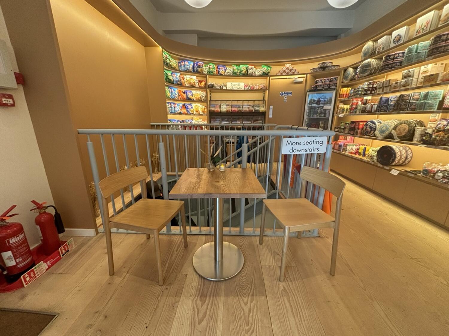 From end of counter facing straight. Table and chairs ahead. Product shelves along left and back walls. Path on right to bathroom at back.