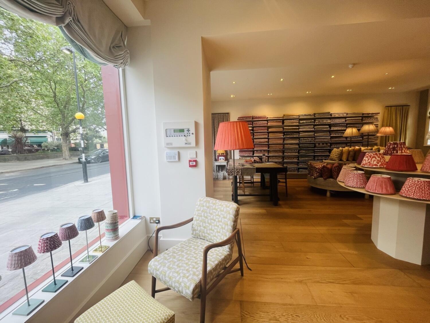 From entrance facing left. Large window with display along left. Chair and lamp in centre. Product shelves along back wall, display counter right.