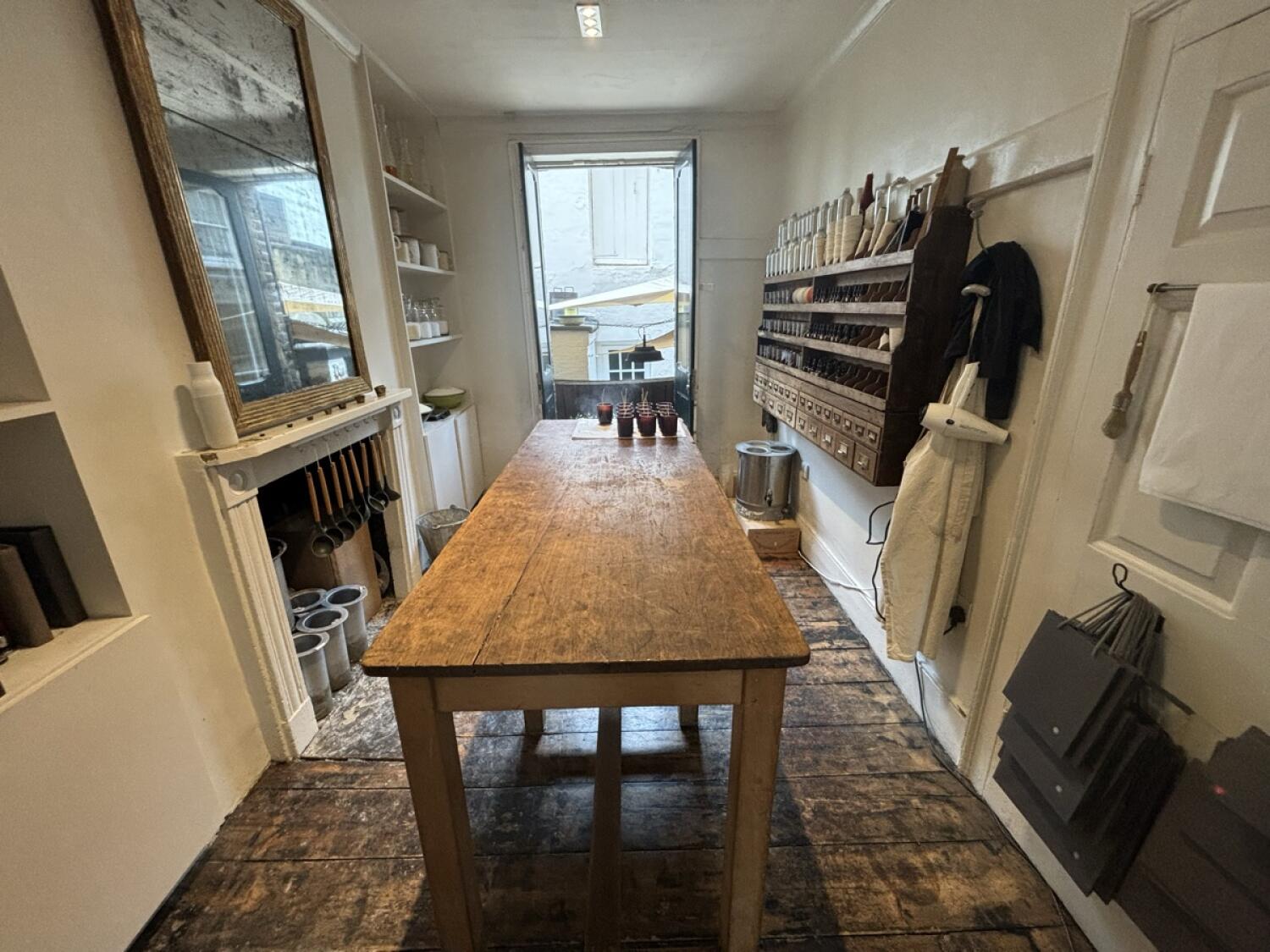 From opening facing straight. Long wooden table in centre. Product shelves along right and left walls. Window at back.