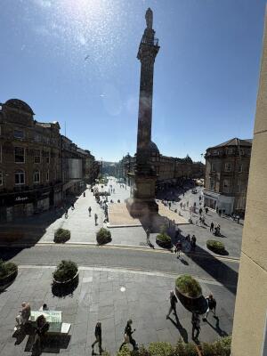 Paved square and buildings left. Monument in centre. Pedestrian street right.