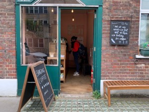 From street, facing entrance. Path straight, between sign on left and outdoor seating on right, to entrance door ahead.