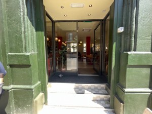 Double glass doors in black metal frame set back from pavement. Silver metal handle in centre. Push to open.